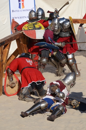A group of armored knights engaged in a medieval-style battle in a sandy arena. They wear shiny metal helmets and chainmail, with bright red tunics over their armor. Some knights have fallen to the ground, while others clash with shields and swords. A banner in the background indicates an event named 'Battle of the Nations'.