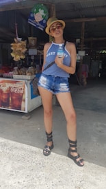A person stands confidently outdoors, wearing a light summer outfit with a blue sleeveless top and distressed denim shorts. They have sunglasses and a straw hat, holding a bottled drink. Behind them is a small shop or stall with signs and snacks on display, including Coca-Cola branding.