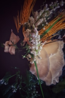 A close-up view of an artistic arrangement of flowers featuring delicate white blossoms, beige petals, and vibrant green foliage. Some elements appear dried with elongated, wheat-like structures providing contrast.