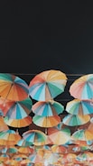 An array of colorful umbrellas is suspended high above the ground, creating a vibrant canopy against a dark background. Each umbrella features a unique combination of bright colors, including shades of blue, red, green, yellow, and orange. The umbrellas are arranged in neat rows and are connected by strings, casting a playful and whimsical vibe.