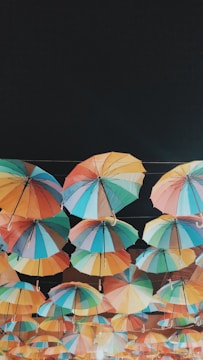An array of colorful umbrellas is suspended high above the ground, creating a vibrant canopy against a dark background. Each umbrella features a unique combination of bright colors, including shades of blue, red, green, yellow, and orange. The umbrellas are arranged in neat rows and are connected by strings, casting a playful and whimsical vibe.