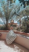 A cozy outdoor patio features a circular seating area with a striped pillow resting on it. Tall palm trees with lush green fronds arch overhead, providing ample shade, while some sunlight creates intricate shadow patterns on the seating. In the background, a serene landscape includes mountains and clear blue sky.