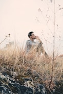 A person is sitting quietly among tall, dry grass in a contemplative posture, hands clasped and raised to their face. The background is a hazy sky with some urban elements faintly visible. The foreground is dominated by rocky terrain with patches of moss.