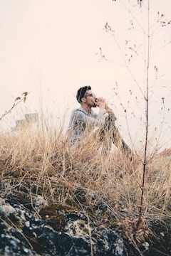 A person is sitting quietly among tall, dry grass in a contemplative posture, hands clasped and raised to their face. The background is a hazy sky with some urban elements faintly visible. The foreground is dominated by rocky terrain with patches of moss.
