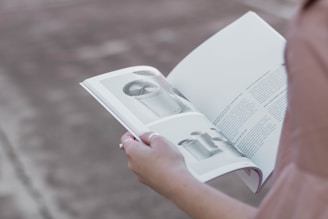 A person holds an open magazine or book, focusing on a page with minimal text and monochrome images of cylindrical objects. The background is blurred, placing emphasis on the reading material.