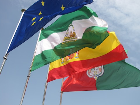 Four flags are waving in the wind against a partly cloudy sky. The flags include the European Union flag with yellow stars on a blue background, a green and white flag with an emblem in the middle, a yellow and red flag with an emblem, and a green and red flag with an emblem.
