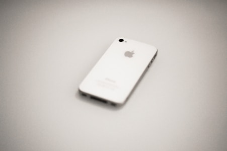 A white smartphone, identified as an Apple iPhone, is placed on a plain, light-colored surface. The phone is oriented with its back facing upwards, featuring the iconic Apple logo in the center. The image is slightly out of focus, providing a soft and minimalistic appearance.