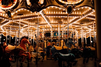 A brightly lit carousel features ornate, colorful horses and is adorned with numerous lights. The carousel's vivid colors contrast with the dark background, creating a warm and inviting ambiance.