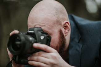 A bald man with a beard is intently looking through the viewfinder of a Canon camera while holding it up close to his face. He appears to be focused on capturing a shot, dressed in a dark suit or coat. The background is blurred, emphasizing his concentration on the photography task.