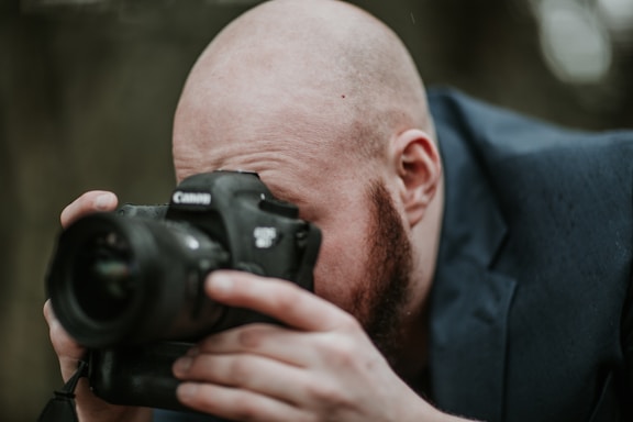 A bald man with a beard is intently looking through the viewfinder of a Canon camera while holding it up close to his face. He appears to be focused on capturing a shot, dressed in a dark suit or coat. The background is blurred, emphasizing his concentration on the photography task.