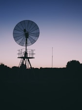 A silhouette of a large satellite dish stands against a gradient sky transitioning from pale pink to deep blue. The structure appears to be set in a rural or remote location, surrounded by trees and open land, suggesting an evening or early morning setting.