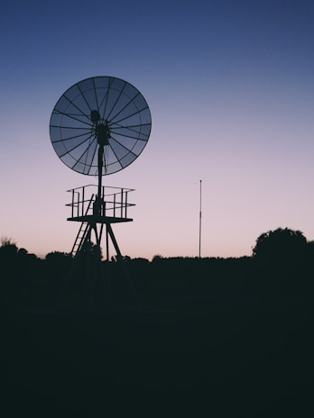 A silhouette of a large satellite dish stands against a gradient sky transitioning from pale pink to deep blue. The structure appears to be set in a rural or remote location, surrounded by trees and open land, suggesting an evening or early morning setting.