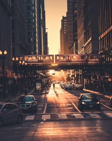 A bustling urban street scene at sunset, with sunlight creating a warm glow over the landscape. Tall buildings line the street, and an elevated train travels across the bridge in the background. Numerous cars are visible, along with pedestrians crossing and walking along the sidewalks.