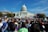 A large crowd gathers in front of a government building with a prominent dome. Many people hold rainbow flags and signs advocating for equality and rights. The blue sky and lush green trees frame the scene, suggesting a peaceful and hopeful demonstration.