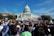 A large crowd gathers in front of a government building with a prominent dome. Many people hold rainbow flags and signs advocating for equality and rights. The blue sky and lush green trees frame the scene, suggesting a peaceful and hopeful demonstration.