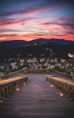 A grand stone staircase descends into a scenic outdoor gathering area with elegantly set tables and chairs. Groups of people are seated in a lush green garden during sunset, with a vibrant sky painted in shades of pink, orange, and purple. The distant hills are silhouetted against the dramatic horizon.