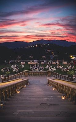 A grand stone staircase descends into a scenic outdoor gathering area with elegantly set tables and chairs. Groups of people are seated in a lush green garden during sunset, with a vibrant sky painted in shades of pink, orange, and purple. The distant hills are silhouetted against the dramatic horizon.