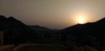 The image captures a serene sunset over a hilly landscape with silhouetted mountains and a valley below. The sun is low on the horizon, casting a warm glow across the scene. There are faint structural outlines suggesting buildings or monuments in the distance.