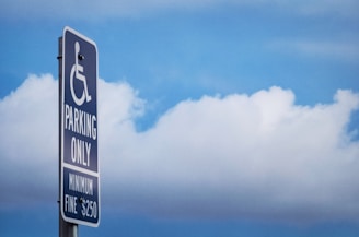 A sign indicating parking reserved for people with disabilities is mounted on a pole against a backdrop of blue sky and clouds. The sign includes the international symbol of access, with text below stating 'Parking Only' and a 'Minimum Fine $250'.