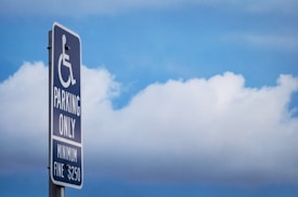 A sign indicating parking reserved for people with disabilities is mounted on a pole against a backdrop of blue sky and clouds. The sign includes the international symbol of access, with text below stating 'Parking Only' and a 'Minimum Fine $250'.