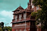 A historical brick building with intricate architecture features a sign that reads 'Baroda Museum & Picture Gallery.' The structure has multiple levels with pointed, tiled roofs and large windows. Surrounded by lush green trees, it sits under a cloudy sky.