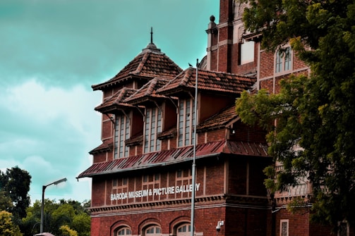 A historical brick building with intricate architecture features a sign that reads 'Baroda Museum & Picture Gallery.' The structure has multiple levels with pointed, tiled roofs and large windows. Surrounded by lush green trees, it sits under a cloudy sky.