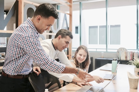 Three people are gathered around a laptop in a modern office setting. They appear to be collaborating on a project, with one person pointing at the laptop screen while the others are looking intently. The environment is well-lit with large windows and there are books and plants on the desk.