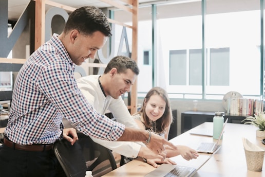 Three people are gathered around a laptop in a modern office setting. They appear to be collaborating on a project, with one person pointing at the laptop screen while the others are looking intently. The environment is well-lit with large windows and there are books and plants on the desk.
