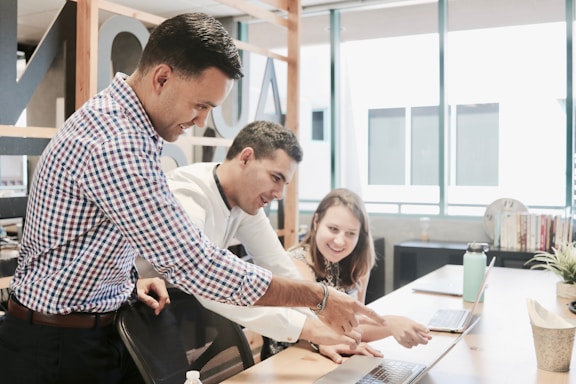 Three people are gathered around a laptop in a modern office setting. They appear to be collaborating on a project, with one person pointing at the laptop screen while the others are looking intently. The environment is well-lit with large windows and there are books and plants on the desk.