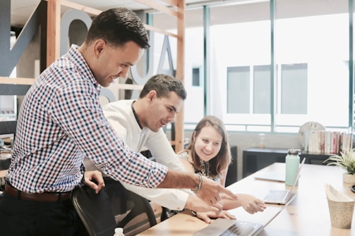 Three people are gathered around a laptop in a modern office setting. They appear to be collaborating on a project, with one person pointing at the laptop screen while the others are looking intently. The environment is well-lit with large windows and there are books and plants on the desk.