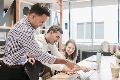 Three people are gathered around a laptop in a modern office setting. They appear to be collaborating on a project, with one person pointing at the laptop screen while the others are looking intently. The environment is well-lit with large windows and there are books and plants on the desk.