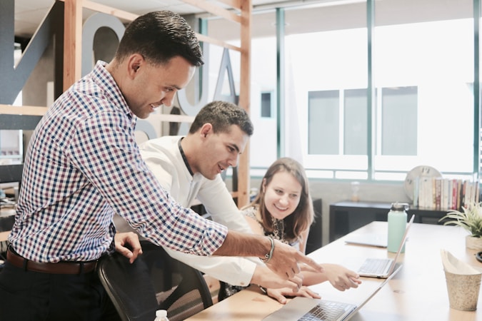 Three people are gathered around a laptop in a modern office setting. They appear to be collaborating on a project, with one person pointing at the laptop screen while the others are looking intently. The environment is well-lit with large windows and there are books and plants on the desk.