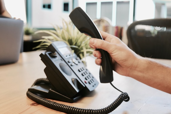 A hand is holding a telephone receiver above a desk. The phone is a traditional office model with a keypad and coiled cord. In the background, there is a blurred view of an office space, including some potted plants and chairs.