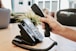 A hand is holding a telephone receiver above a desk. The phone is a traditional office model with a keypad and coiled cord. In the background, there is a blurred view of an office space, including some potted plants and chairs.