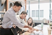 Three people are collaborating in a bright office space, focused on a laptop screen. The atmosphere suggests teamwork and discussion as one person points at the laptop while the others pay attention.