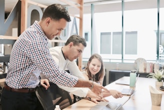 Three people are collaborating in a bright office space, focused on a laptop screen. The atmosphere suggests teamwork and discussion as one person points at the laptop while the others pay attention.