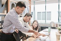 Three people are collaborating in a bright office space, focused on a laptop screen. The atmosphere suggests teamwork and discussion as one person points at the laptop while the others pay attention.