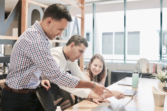 Three people are collaborating in a bright office space, focused on a laptop screen. The atmosphere suggests teamwork and discussion as one person points at the laptop while the others pay attention.