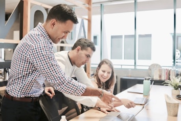 Three people are collaborating in a bright office space, focused on a laptop screen. The atmosphere suggests teamwork and discussion as one person points at the laptop while the others pay attention.