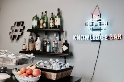 The image features a wall with a neon sign reading 'Knowledge Bar,' which is shaped like an apple with glasses. Below the sign, there are shelves filled with various bottles of liquor. In the foreground, a table is set up with a bowl of fresh fruit including oranges, bananas, and apples, alongside a tray filled with bottled water and wrapped food items. A decorative hashtag symbol is mounted on the wall beside the shelves.
