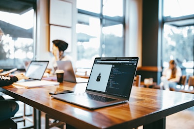 Laptops placed on a wooden table in a modern cafe setting with people in the background working or relaxing. Natural light comes through large windows, creating a bright and airy atmosphere.