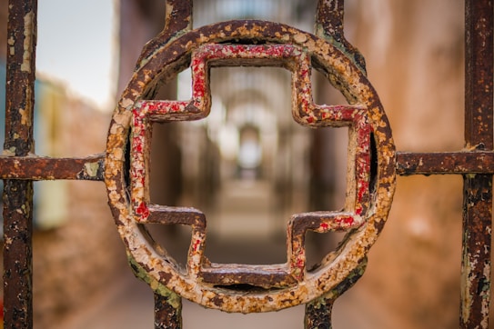 A rusted metal gate featuring a circular design at its center, heavily textured with peeling paint in red and creamy beige hues. The background is blurred, creating a sense of depth and focus on the gate structure.