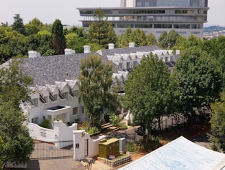 A large, elegant white building with a slate roof situated amidst dense greenery. The architecture features multiple gabled dormers and a gated entrance, adding to the stately appearance. In the background, a modern multi-story building is visible, contrasting with the classic style of the foreground structure.