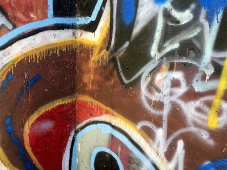 The image features a wall covered in vibrant graffiti with abstract shapes and lines. Prominent colors include red, blue, and yellow with black outlines and white highlights, creating a dynamic and energetic composition. Drips of paint are visible, adding to the urban art feel.