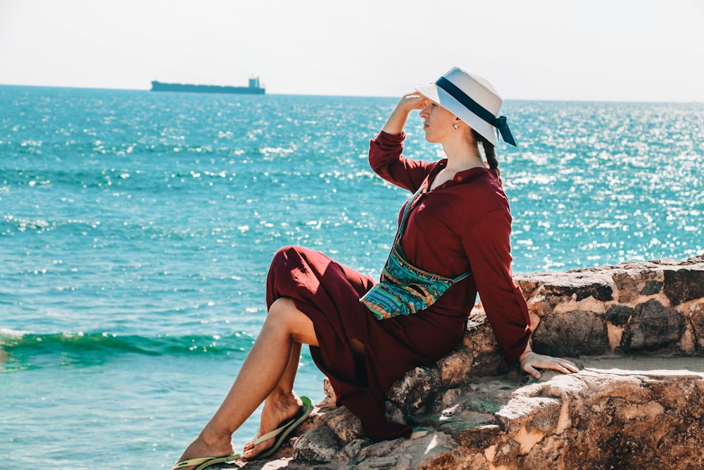 Mujer sentada en la roca y poniendo la mano derecha sobre su cabeza viendo el mar en calma