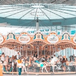 A colorful carousel indoors with ornate decorations and paintings set against a modern architectural backdrop. Several people, including adults and children, are riding the carousel horses, enjoying the experience.