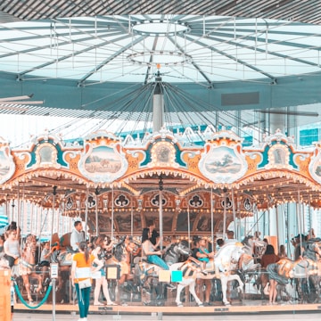A colorful carousel indoors with ornate decorations and paintings set against a modern architectural backdrop. Several people, including adults and children, are riding the carousel horses, enjoying the experience.