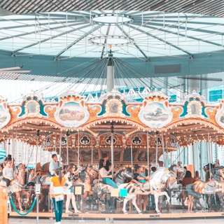 A colorful carousel indoors with ornate decorations and paintings set against a modern architectural backdrop. Several people, including adults and children, are riding the carousel horses, enjoying the experience.