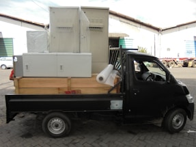A black truck parked in an industrial area is loaded with large metal cabinets and a roll of white material. The truck's bed is wooden and surrounded by various parked vehicles in a warehouse environment.
