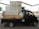 A black truck parked in an industrial area is loaded with large metal cabinets and a roll of white material. The truck's bed is wooden and surrounded by various parked vehicles in a warehouse environment.